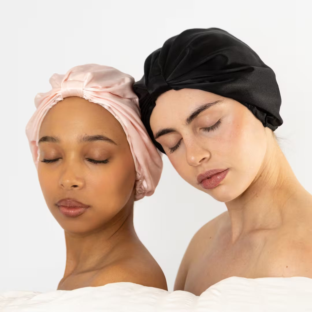Two women resting together, one in a blush pink and one in an onyx black organic silk bonnet